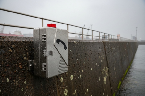 Silver weatherproof emergency phone enclosure mounted on seawall in rainy coastal environment