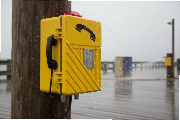Yellow weatherproof SIP hotline phone on wooden post, rain splash at seaport