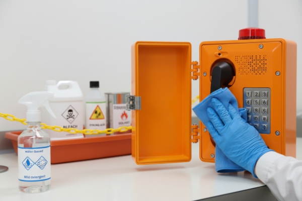 Gloved hand wiping orange industrial SIP emergency phone near labeled chemical containers