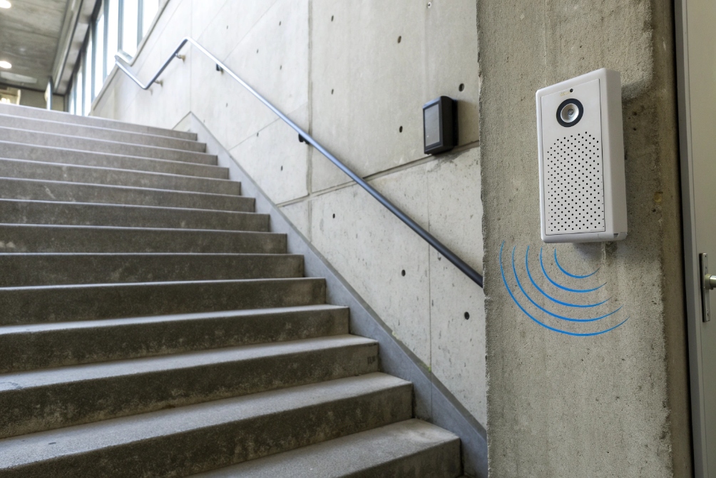 White wall-mounted intercom with camera and speaker on a concrete wall beside a stairwell, with blue sound-wave graphics indicating audio transmission.