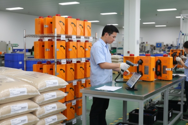 Quality inspector testing orange SIP emergency phones on assembly line with stock racks