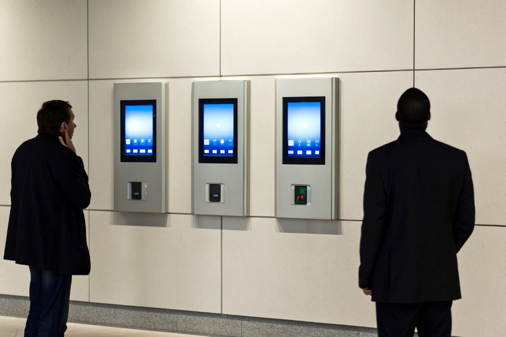 Two people standing in lobby in front of three large wall-mounted touchscreen access terminals